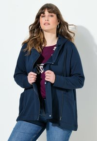 Sudadera con cremallera de color azul marino, con capucha, dos bolsillos frontales y un corte relajado, llevada sobre una camiseta burdeos con un diseño ligero.