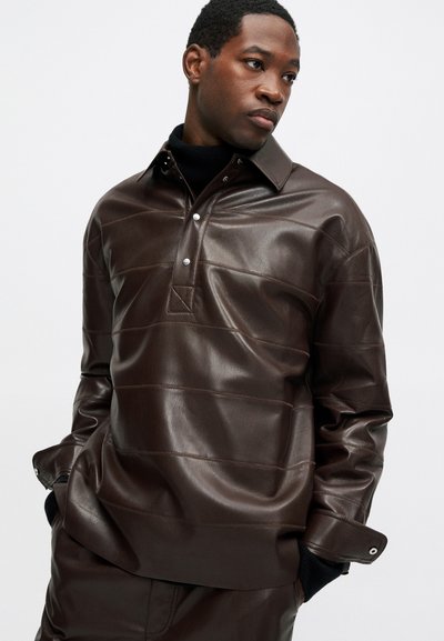 Férfi sötétbarna bőr ingben és hozzá illő nadrágban, kezében a zsebében, az oldalra néz egy sima háttér előtt.