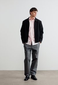 Άνδρας που στέκεται με τα χέρια στις τσέπες, φοράει μαύρο μπουφάν, ανοιχτό ροζ πουκάμισο, γκρι παντελόνι και μαύρα παπούτσια πάνω σε απλό λευκό φόντο.