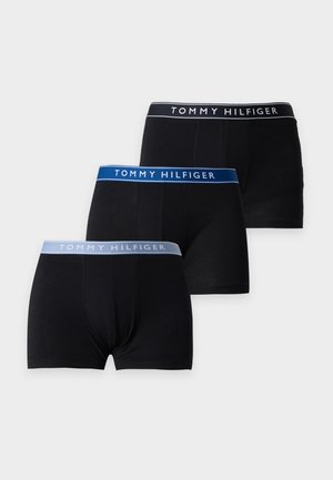 Homme athlétique debout pieds nus, portant un boxer noir Tommy Hilfiger avec une ceinture bleu clair sur un fond uni clair.