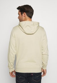 Beige hettegenser laget av myk stoff, med kenguru-lomme, ribbestrikkede mansjetter og søm. Båret med mørke denimjeans.