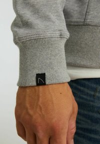 Puño de sudadera gris con textura acanalada, que presenta una pequeña etiqueta negra con un diseño de contorno blanco. Mano parcialmente visible con detalle de textura de piel.