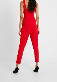 Mono rojo con tirantes anchos, un corsé ajustado y piernas estrechas. Textura suave y diseño minimalista. Combinado con tacones altos negros.
