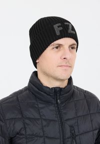 Svart stickad mössa med ribbat mönster och grå "FZ"-bokstäver. Bärs av en person i en svart zip-up pufferjacka.