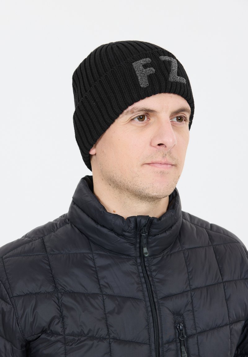 Svart stickad mössa med ribbat mönster och grå "FZ"-bokstäver. Bärs av en person i en svart zip-up pufferjacka.
