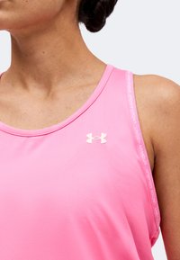 Γυναίκα που φοράει ροζ αμάνικο τοπ Under Armour με λογότυπο στο στήθος και λουράκι με σήμα στον ώμο, μπροστά σε απλό φόντο.