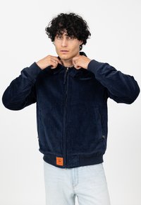 Marineblauwe ribfluwelen ritsjack met ribgebreide manchetten en zoom, voorzien van zijzakken en een oranje merklabel aan de onderkant.