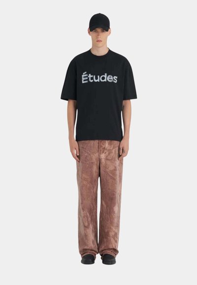 Jeune homme portant une casquette noire, un t-shirt noir "Études" et un pantalon ample marron texturé, debout devant un fond clair uni.