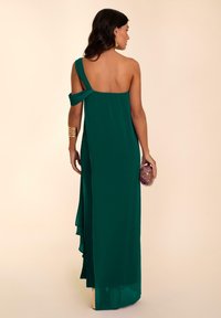 Langes, asymmetrisches Kleid in Smaragdgrün, mit einem schmalen Träger, fließendem Stoff und einem seitlichen Schlitz. Akzentuiert mit einer strukturierten Clutch.