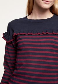 Meriharmaa ja burgundy raidoitettu neulepaita, jossa on ribattu kaula-aukko ja röyhelökoristus olkapäässä. Pehmeä neulemateriaali, kapea istuvuus.