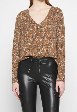 Kobieta ubrana w brązową kwiecistą bluzkę z dekoltem w serek, połączoną z czarnymi, wysokim stanem spodniami z eko skóry z troczkiem w talii.