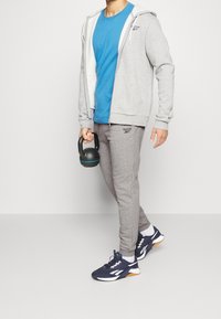 Grå zip-up hoodie över en blå T-shirt, kombinerad med grå sweatpants och marinblå sneakers. Håller en svart kettlebell med turkosa detaljer.