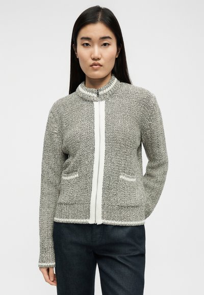 Νεαρή γυναίκα με μακριά σκούρα μαλλιά φοράει μια υφασμάτινη γκρι ζακέτα με φερμουάρ και σκούρο παντελόνι, όρθια μπροστά σε ένα απλό φωτεινό φόντο.