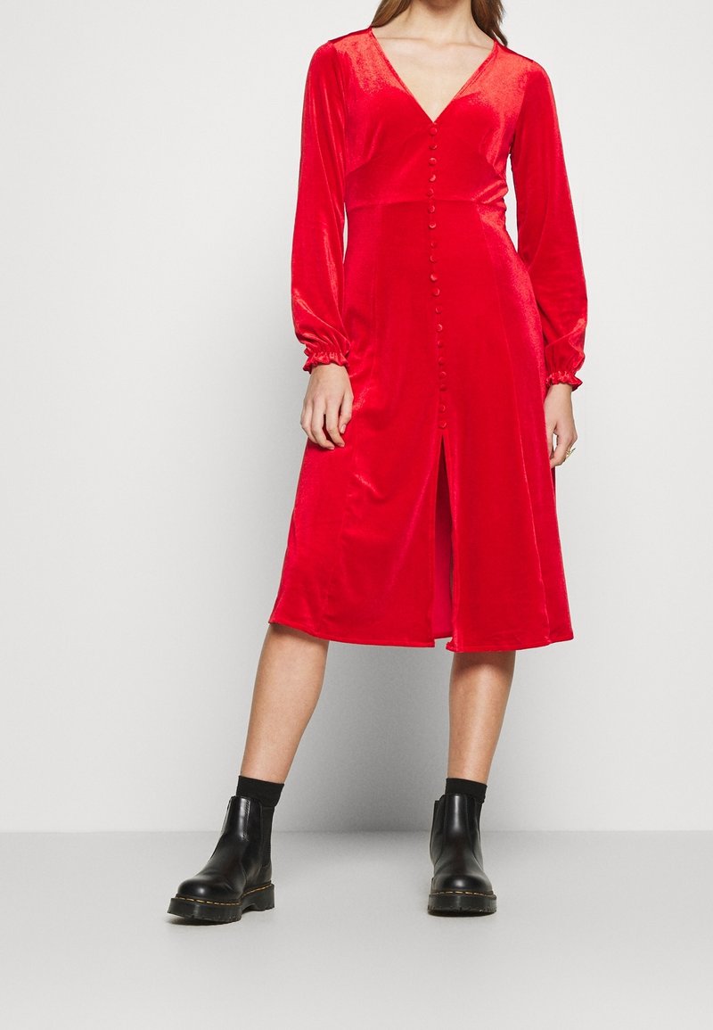 Röd sammet midi-klänning med V-ringning, långa puffärmar och knäppning framtill. Matchas med svarta ankelstövlar. Slät textur, knä-lång nederkant.