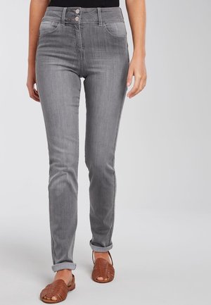 Frau trägt graue, hoch taillierte Jeans mit umgeschlagenen Säumen und braune, gewebte Leder-Sandalen mit offenen Zehen, steht vor einem neutralen Hintergrund.