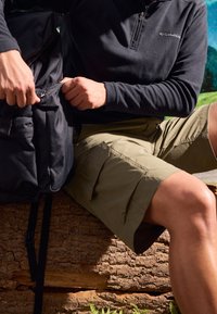 Musta vetoketjullinen reppu ja tumma fleece-paita sekä oliivinvihreät cargo-shortit peittävä henkilö, istu lamekkeella, taustalla ruohoinen maisema.