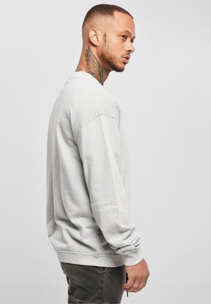 Homme avec des tatouages sur le cou portant un sweat-shirt blanc et un pantalon foncé, debout de profil sur un fond clair uni.