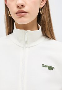 Κοντινό πλάνο ενός ατόμου που φοράει άσπρο μπουφάν με φερμουάρ και λογότυπο Lacoste, καθώς και χρυσούς κρίκους σκουλαρίκια, εμφανίζοντας τον λαιμό και το κάτω μέρος του προσώπου.