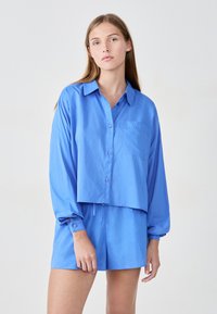 Krótka niebieska koszula z guzikami z przodu i kieszonką na piersi, w połączeniu z dopasowanymi szortami. Oba elementy mają gładką fakturę i luźny krój.