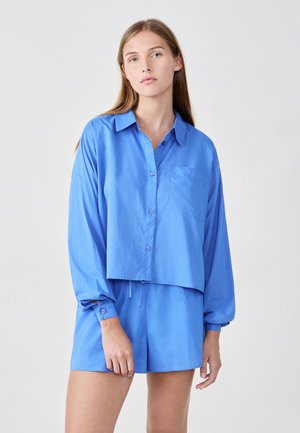 Abgeschnittenes blaues Hemd mit Knopfleiste und Brusttasche, kombiniert mit passenden Shorts. Beide Kleidungsstücke haben eine glatte Textur und eine lockere Passform.