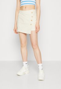 Une mini-jupe de couleur crème avec un design enveloppant, dotée de quatre boutons sur le côté et fabriquée dans un tissu léger.