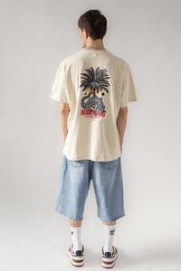 Beiges Baumwoll-T-Shirt mit einem Grafikmotiv eines Palmbaums und einer Schlange auf dem Rücken, kombiniert mit hellblauen Jeansshorts und weißen Socken mit Muster.