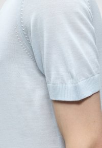 Nahaufnahme eines hellblauen gestrickten Kurzarmkleidungsstücks, das die Stoffstruktur und den gerippten Bündchen am Oberarm zeigt.