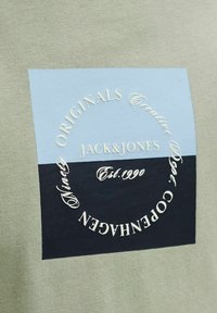 T-shirt en coton vert avec un patch rectangulaire bleu et noir portant un texte blanc, incluant "JACK&JONES" et "COPENHAGEN".