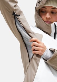 Wasserdichte Jacke in Beige und Weiß. Verfügt über eine Reißverschlusstasche, ein weiches Obermaterial, eine verstellbare Kapuze und ein inneres Mesh-Futter für Belüftung.
