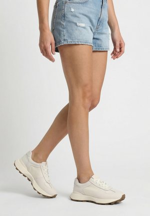 Person, die hellblaue Jeansshorts und weiße Turnschuhe mit braunen Sohlen trägt und vor einem schlichten grauen Hintergrund steht.