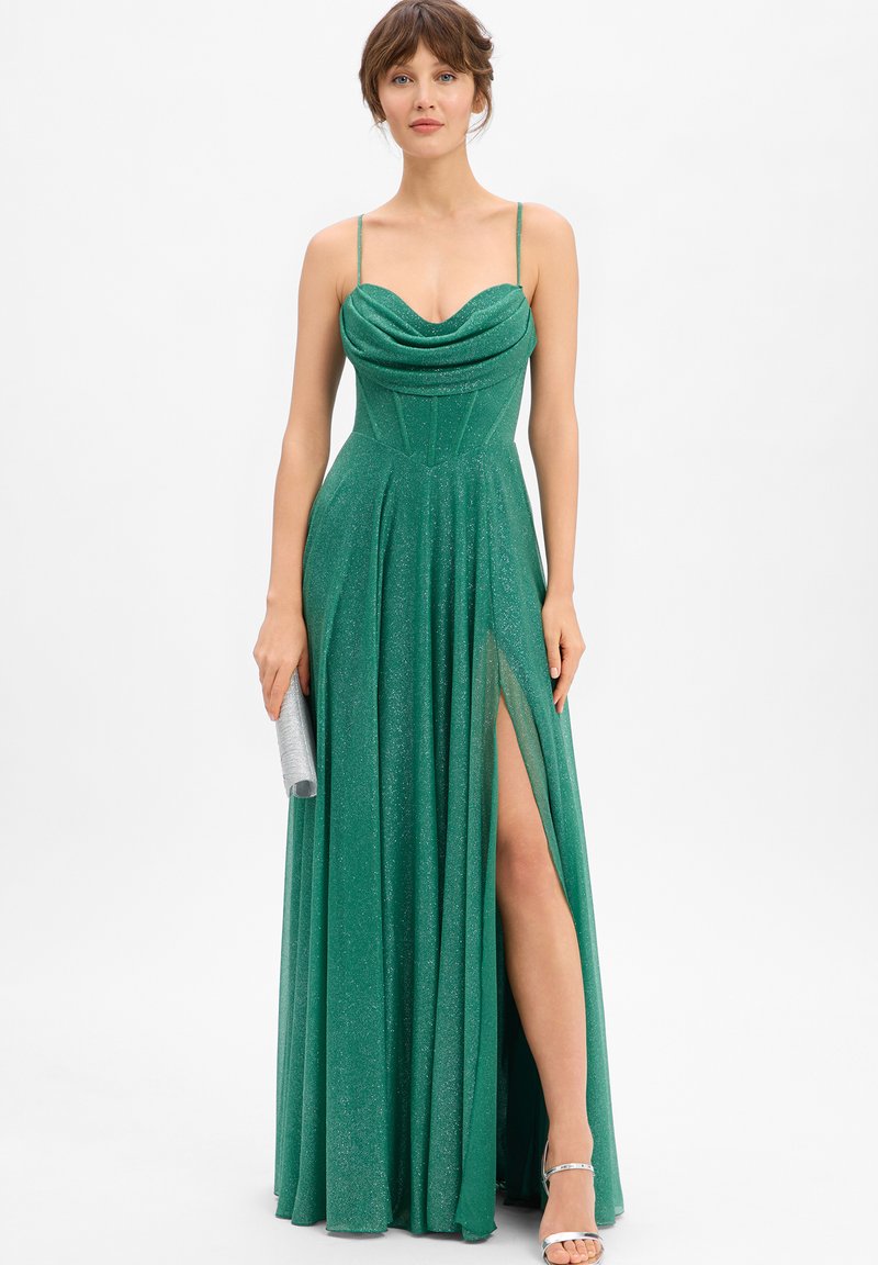 Vrouw die een sprankelende groene avondjurk draagt zonder mouwen met een hoge split tot aan de dij, ze houdt een zilveren clutch vast en draagt zilveren hakken.