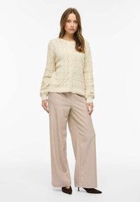 Femme portant un cardigan en tricot crème boutonné avec un motif en diamant, un pantalon beige à jambes larges et des talons noirs pointus, debout devant un fond blanc.