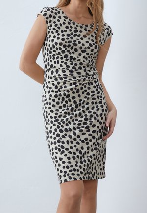 Donna che indossa un vestito beige al ginocchio con pois neri irregolari, in piedi con una mano dietro e l'altra appoggiata i fianchi.