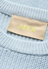 Suéter de malha azul claro com tecido texturizado. Etiqueta apresenta "JJXX" em amarelo sobre um fundo bege. Close-up na área do pescoço.