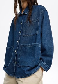 Camicia di jeans blu scuro con colletto, chiusura frontale con bottoni, due grandi tasche anteriori e vestibilità comoda con cuciture in vista.