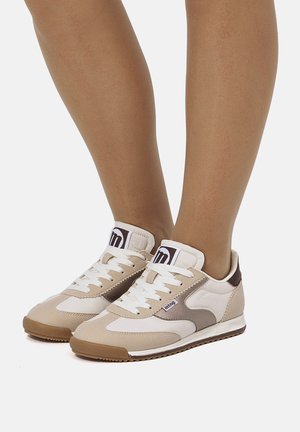Primer plano de piernas con zapatillas de estilo retro en beige, blanco y marrón, con cordones blancos y suela de goma, de pie sobre una superficie blanca.