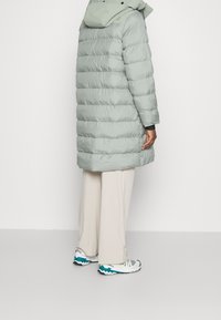 Ljusgrön pufferjacka med horisontell sömnad, en huva och långa ärmar. Buren över krämfärgade byxor och matchad med sneakers med turkosa detaljer.