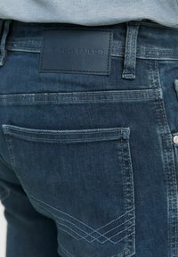 Donkerblauwe denim jeans met een leren label met de opdruk "TOM TAILOR," voorzien van twee achterzakken en decoratieve stiksels in een kruislings patroon.