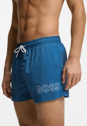 Wysportowany męski tors w niebieskich kąpielówkach z białym sznurkiem i logo "BOSS" na lewej udzie na białym tle.