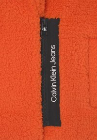Pomarańczowa polarowa kurtka z teksturowanym materiałem i czarnym zamkiem. Zamek ma białe napisy "Calvin Klein Jeans" wzdłuż lewej strony.