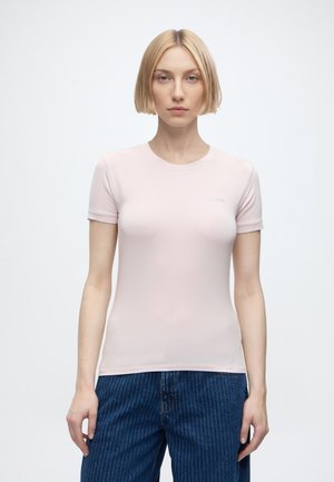 Femme blonde aux cheveux courts portant un t-shirt ajusté rose clair et un jean bleu à fines rayures, debout devant un fond uni.