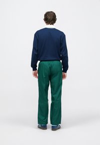 Μπλε σκούρο μπλουζάκι με μακριά μανίκια και λευκό γιακά, συνδυασμένο με άνετο, πράσινο παντελόνι. Το μοντέλο φορά μπλε παπούτσια με ρίγες. Μινιμαλιστικός σχεδιασμός.