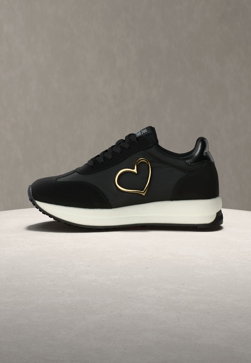 Μαύρο αθλητικό παπούτσι με λεία υφή, που φέρει ένα χρυσό διακοσμητικό καρδιάς στο πλάι, στρογγυλή μύτη και λευκή σόλα. Κορδόνια στο ίδιο μαύρο χ ρώμα.