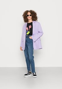 Blazer oversized lilás, t-shirt gráfica preta com estampa colorida, jeans azuis de cintura alta e ténis pretos com solas brancas.