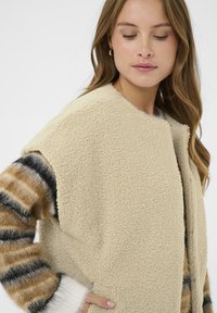 Bežni flis prsluk s teksturiranom površinom, koji ima okrugli izrez, bez rukava i nosi se preko prugastog, višebojnog pulovera.