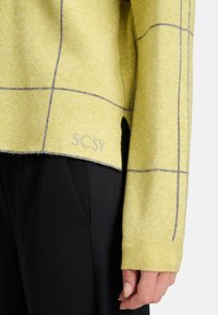 Żółty sweter o puszystej teksturze, z szarymi kratkami i wyraźnym wykończeniem z logo "SCSY". Połączony z czarnymi spodniami.