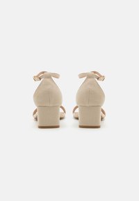 Beige mokka-nahka solkikengät, joissa on takana nilkkavyö, näyttävät sileältä ja minimalistiselta muotoilultaan. Vyössä on kullanvärisiä yksityiskohtia.
