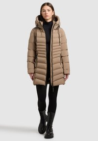 Tan puffersjakke med hætte, der har et quiltet design, lynlås foran og snorede detaljer. Sammen med sorte leggings og kampstøvler.