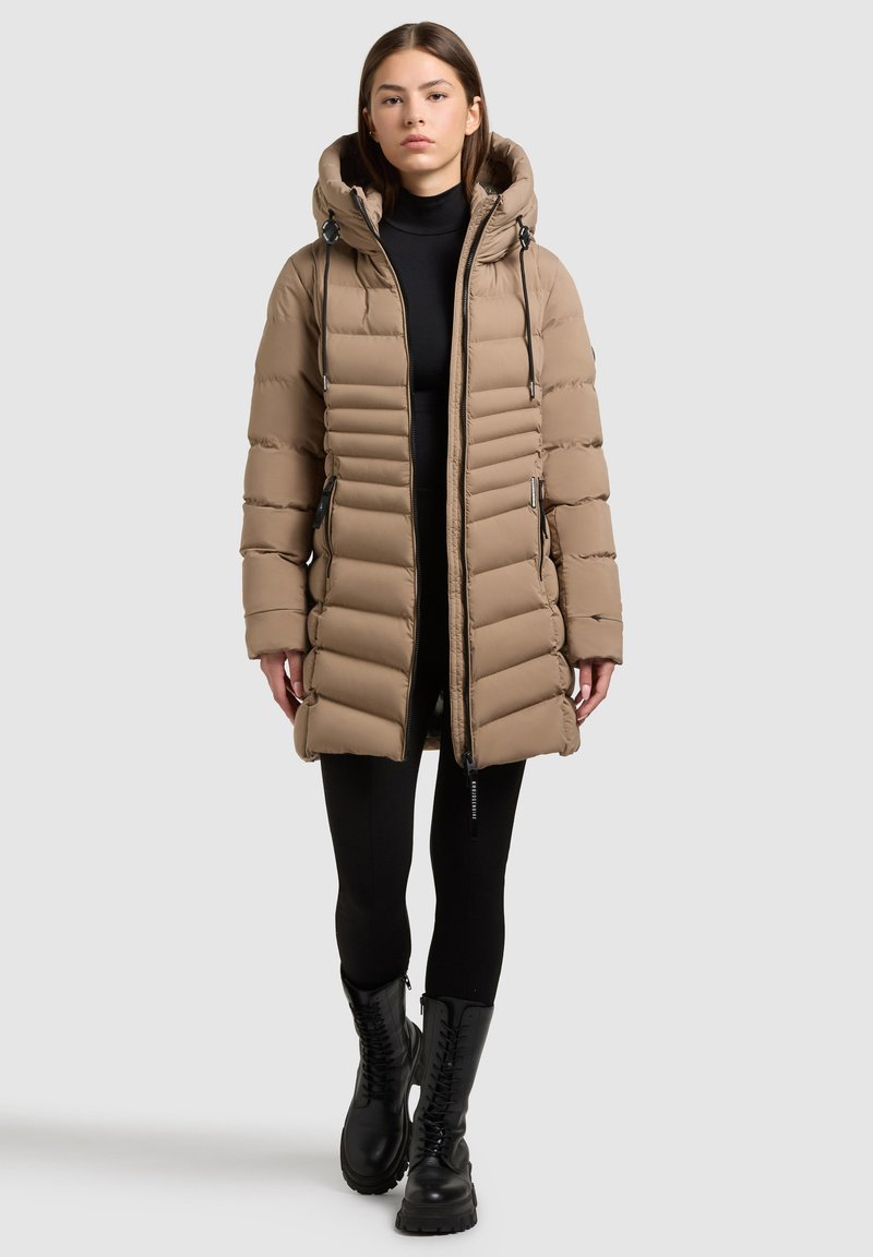Tan puffersjakke med hætte, der har et quiltet design, lynlås foran og snorede detaljer. Sammen med sorte leggings og kampstøvler.