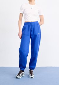 Modré tepláky s elastickými manžetami a šňůrkou v pase, spárované s přiléhavým bílým tričkem s malým černým logem Nike. Tenisky s vzorovaným designem.